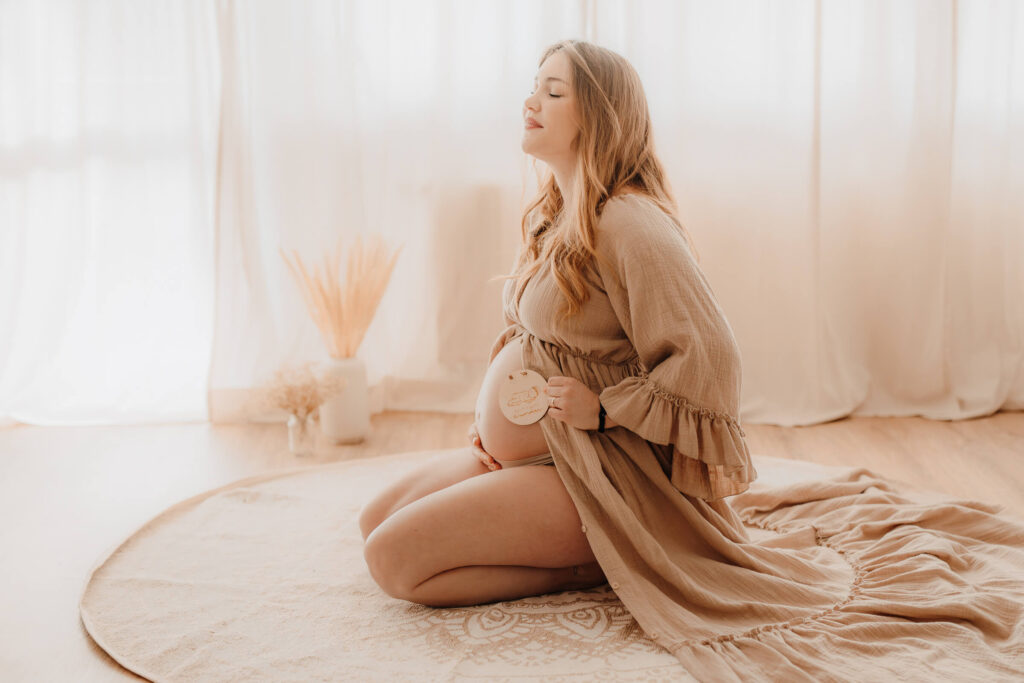 babybauchshooting zuhause, schwangere beruehrt ihren bauch und haelt ein holzschild in der hand