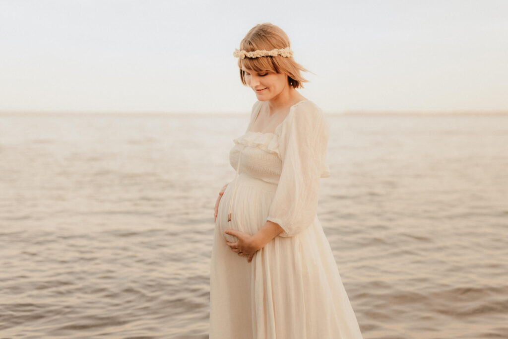 babybauchshooting ostsee strand, schwangere steht im wasser und haelt babybauch fest