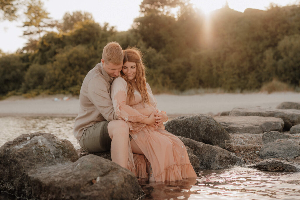 babybauchshooting luebecker bucht, schwangere und partner sitzen auf felsen im wasser