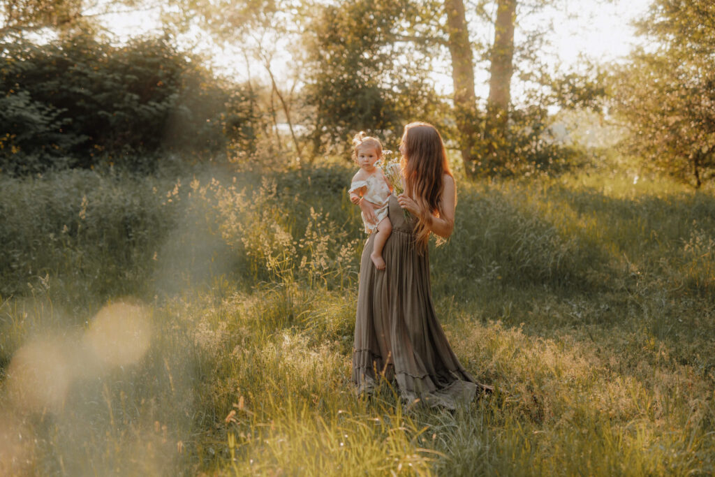 mama kind shooting, mama hat kind auf dem arm, stehen im hohen gras im sonnenuntergang und gucken sich blumen an