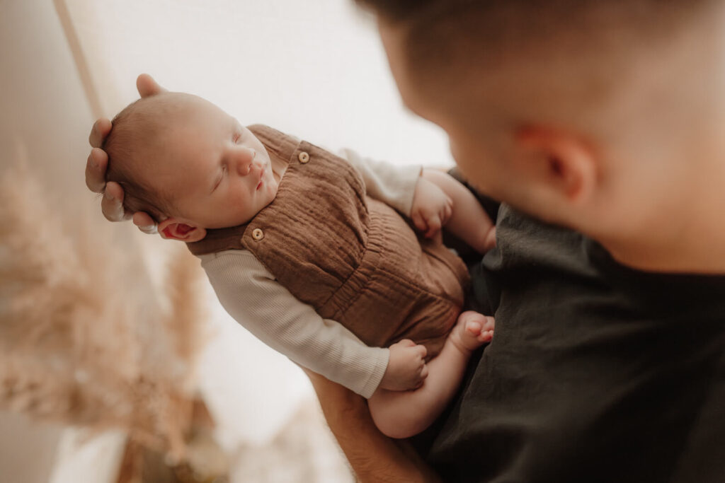 newbornshooting, baby liegt in papas armen und schlaeft, papa guckt es an