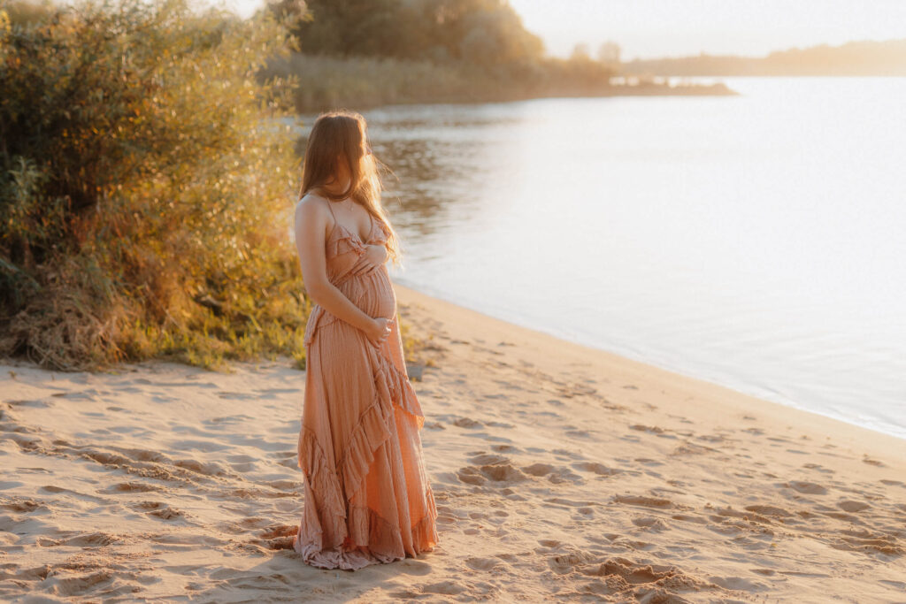 babybauchshooting drage strand, schwangere steht am wasser im sonnenuntergang golden hour